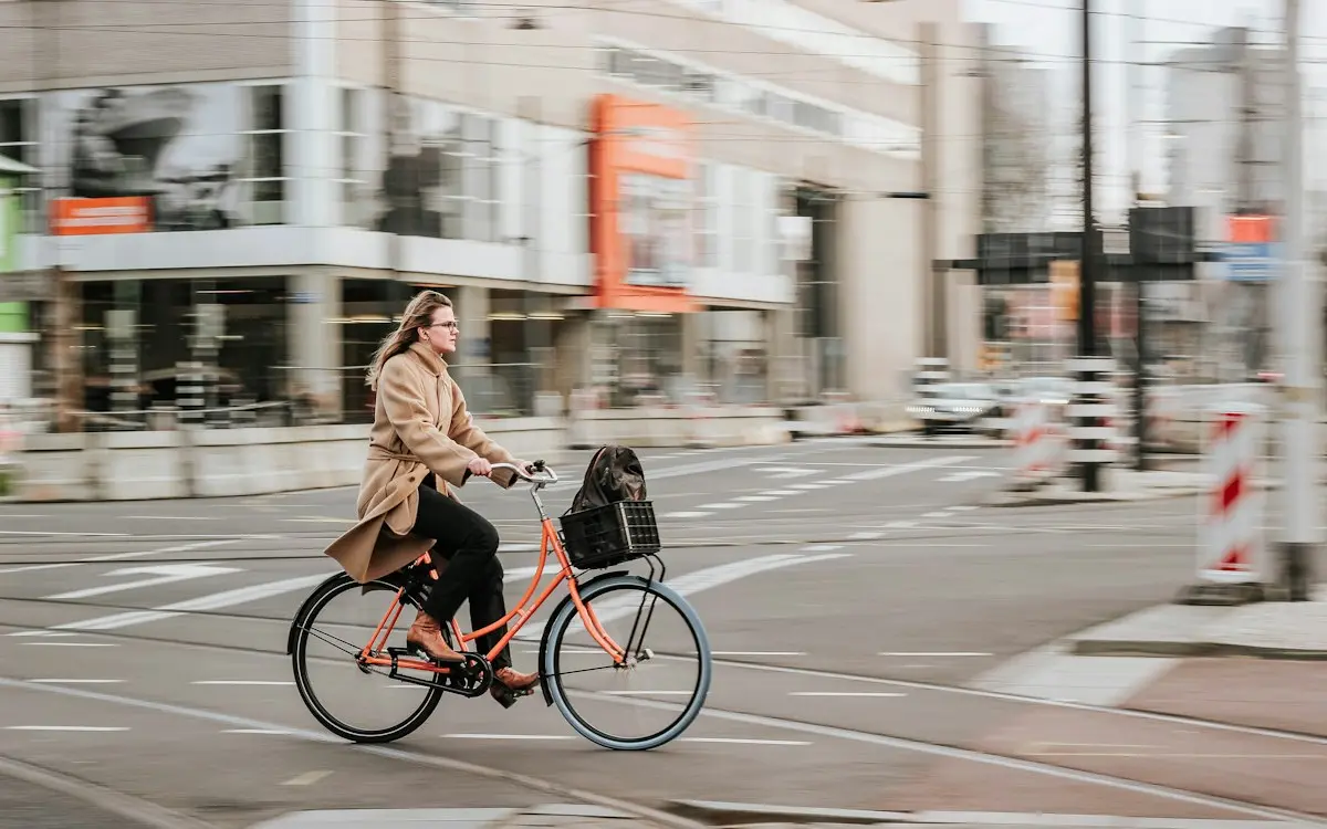 Woman in a camel coat riding an orange city bike with a front basket across a city street, motion-blurred background