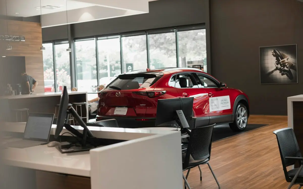 Red SUV on display inside a car dealership showroom, seen past a sales desk with computer monitors