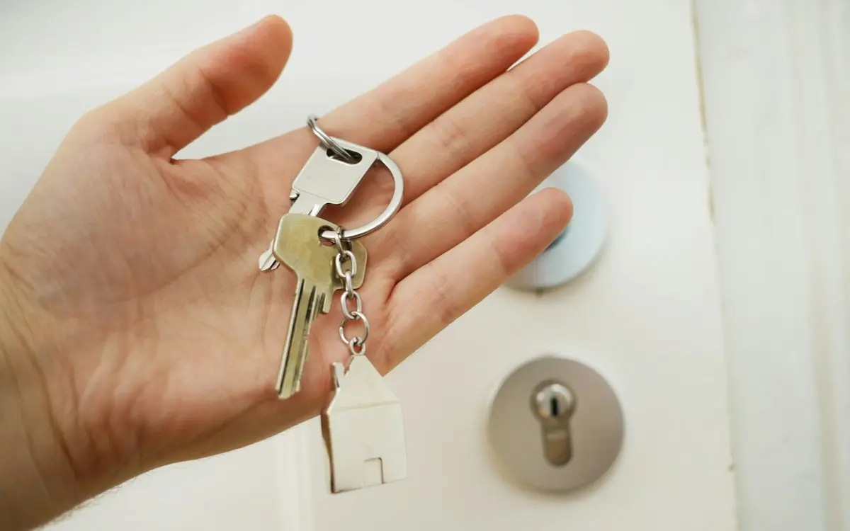 A hand holding a set of apartment keys with a small house-shaped keyring in front of a white door lock