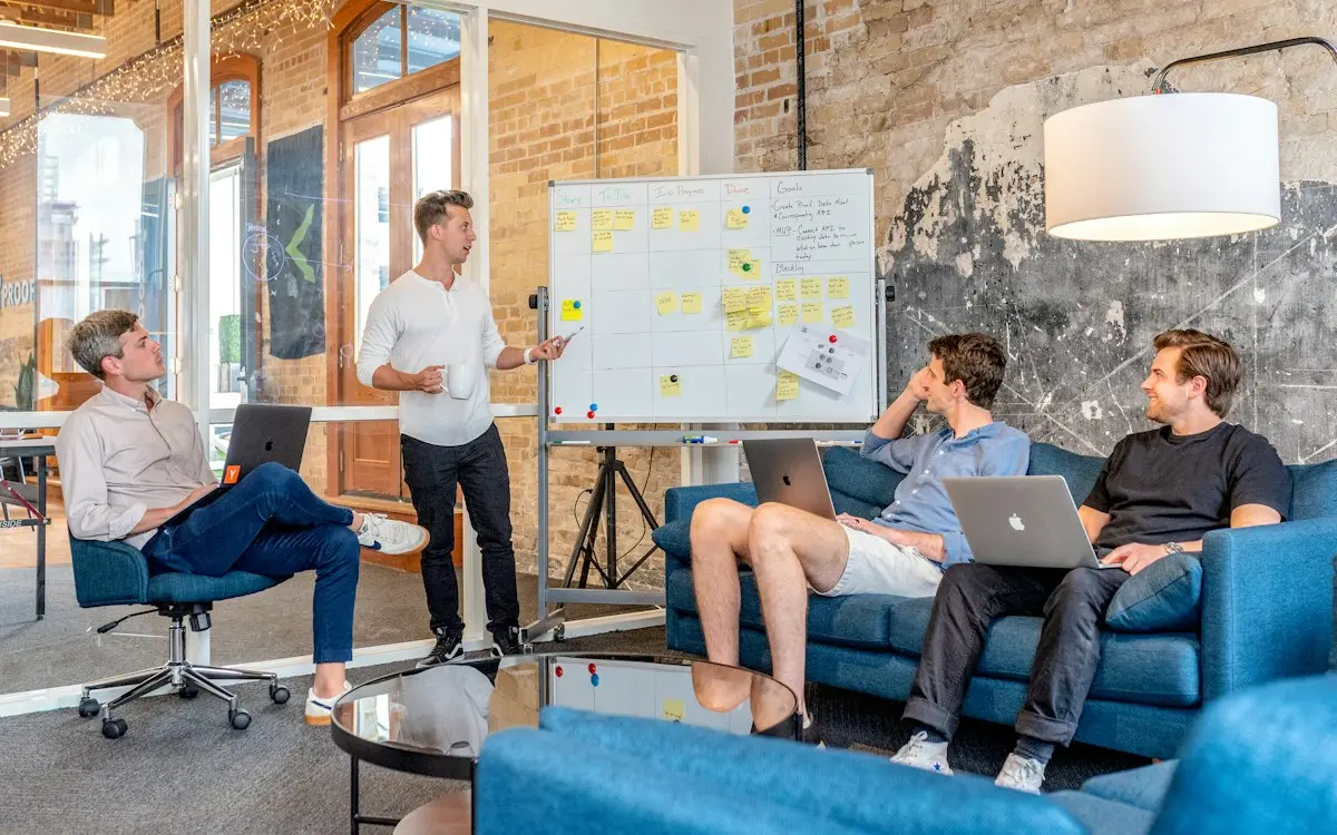 Four young founders in a brick-walled office: one stands at a whiteboard covered in sticky notes while three colleagues sit on blue sofas with laptops